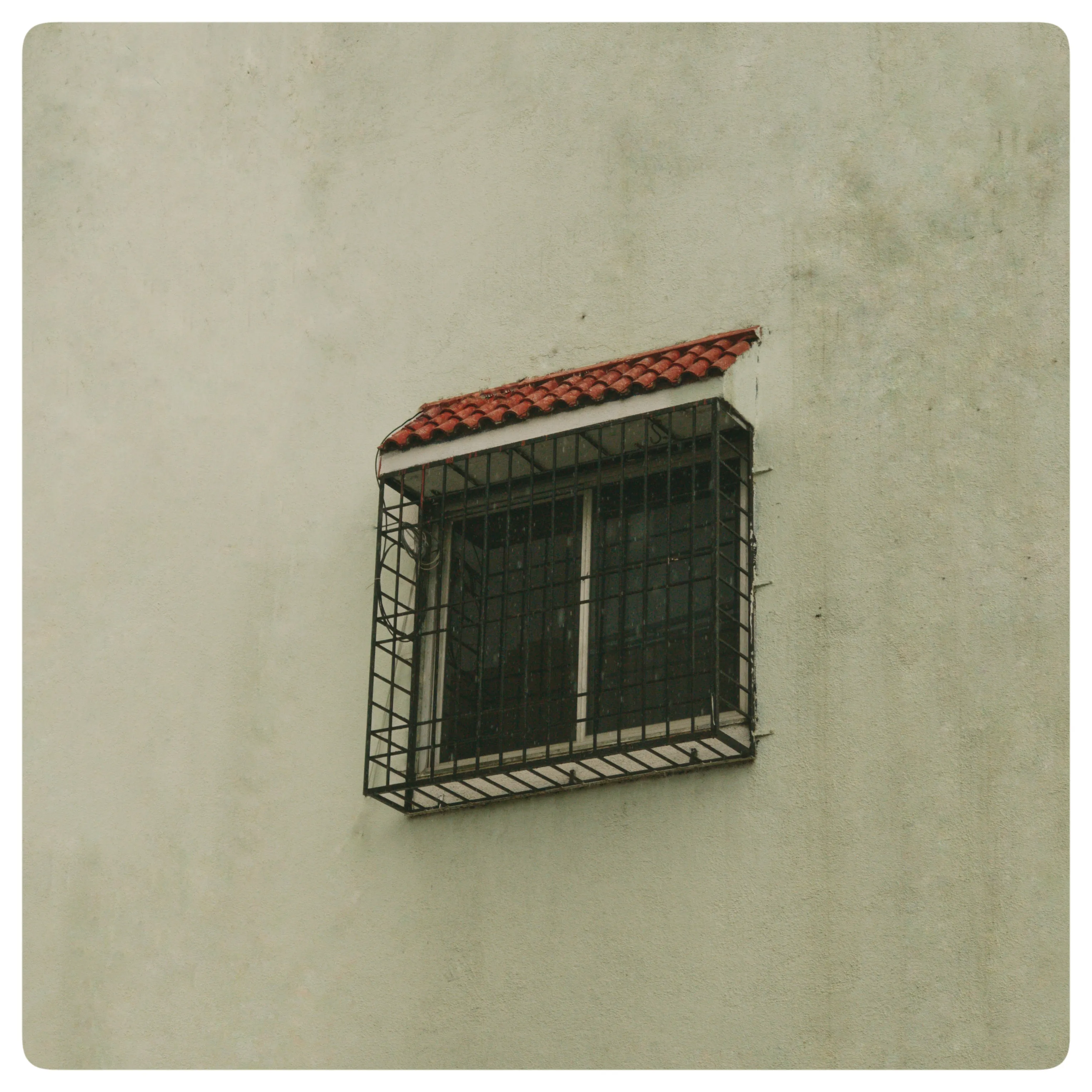 A barred window with a small red-tiled awning on a pale textured wall.