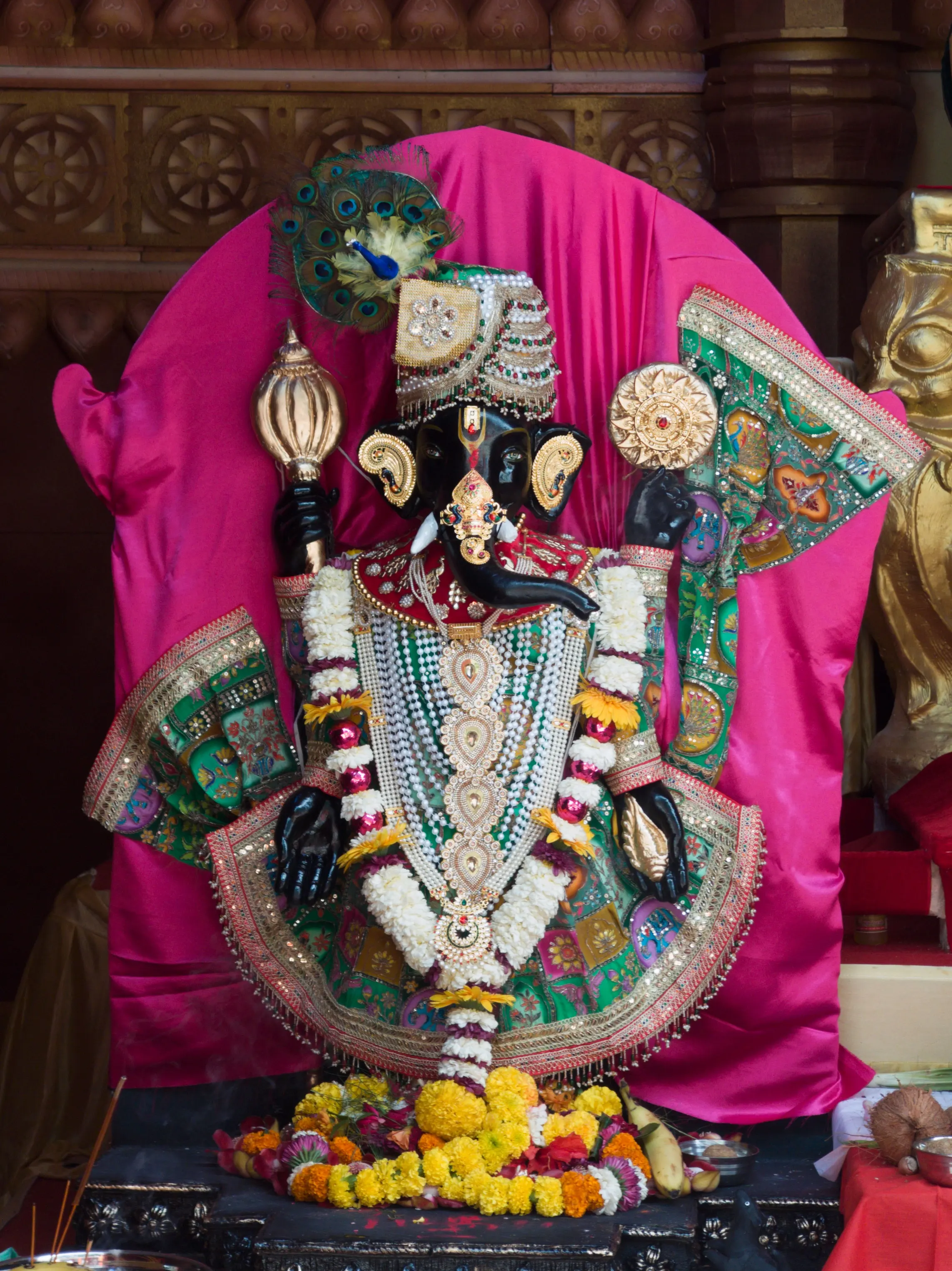 A black Ganesha idol adorned with jewellery and garlands against bright pink drapery.