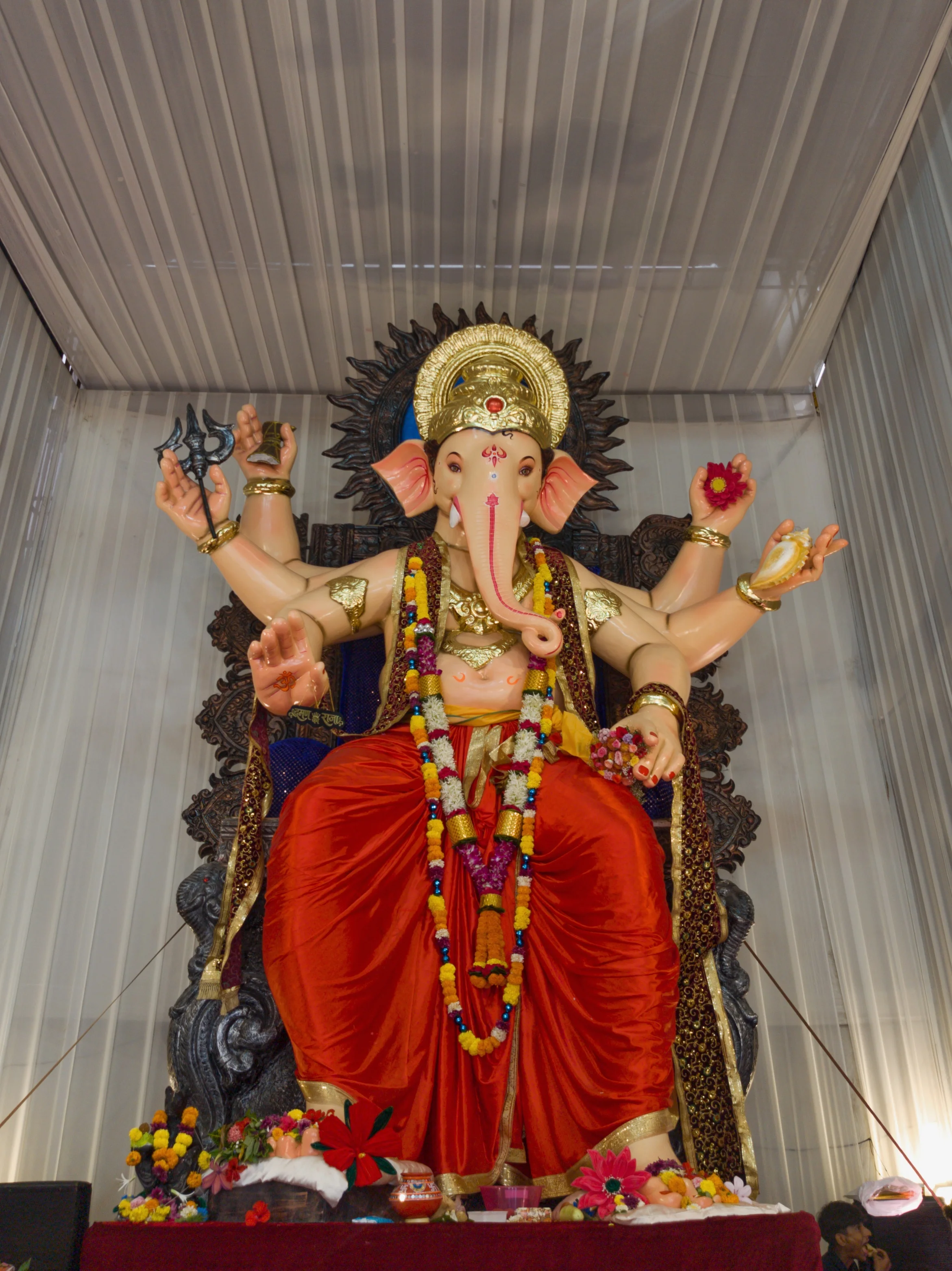 A standing Ganesha idol with a gold crown and red dhoti beneath a corrugated roof.