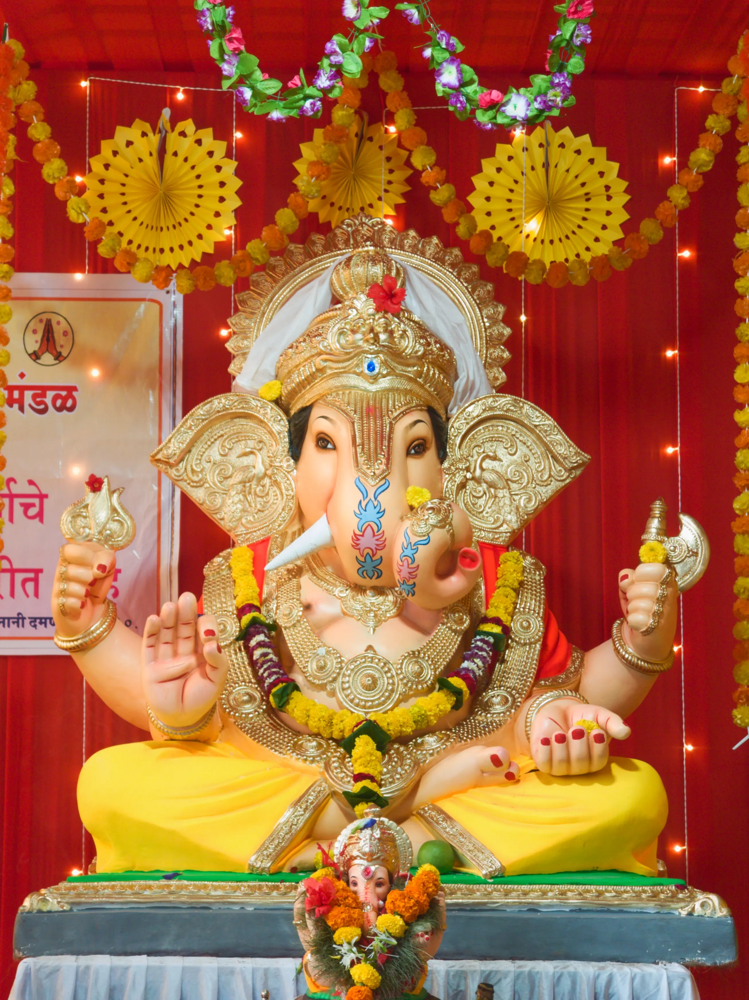 A Ganesha idol with a silver crown and flower garlands sits before a red floral backdrop.