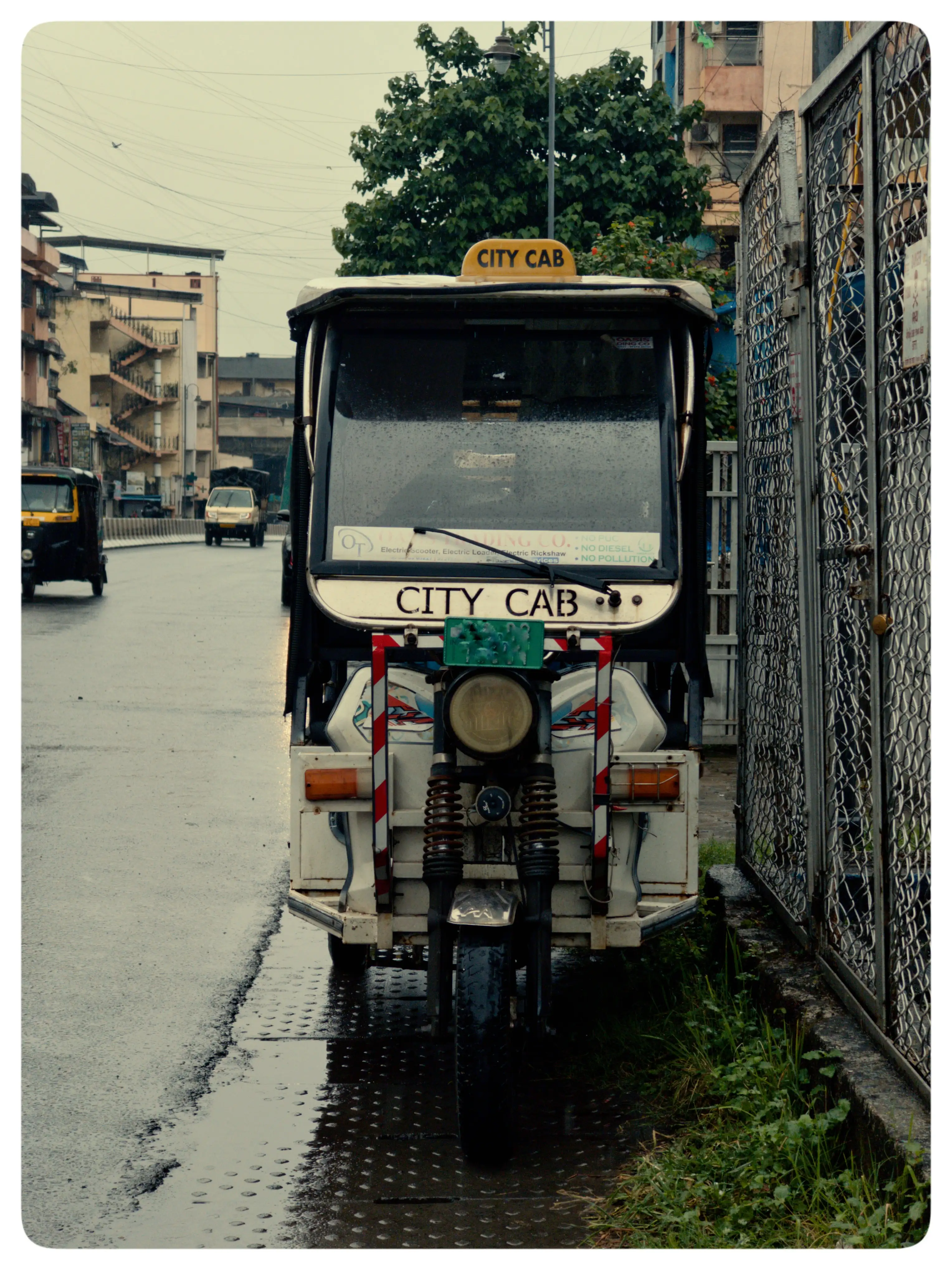 A dark auto-rickshaw stands along a neighbourhood street lined with buildings and trees.